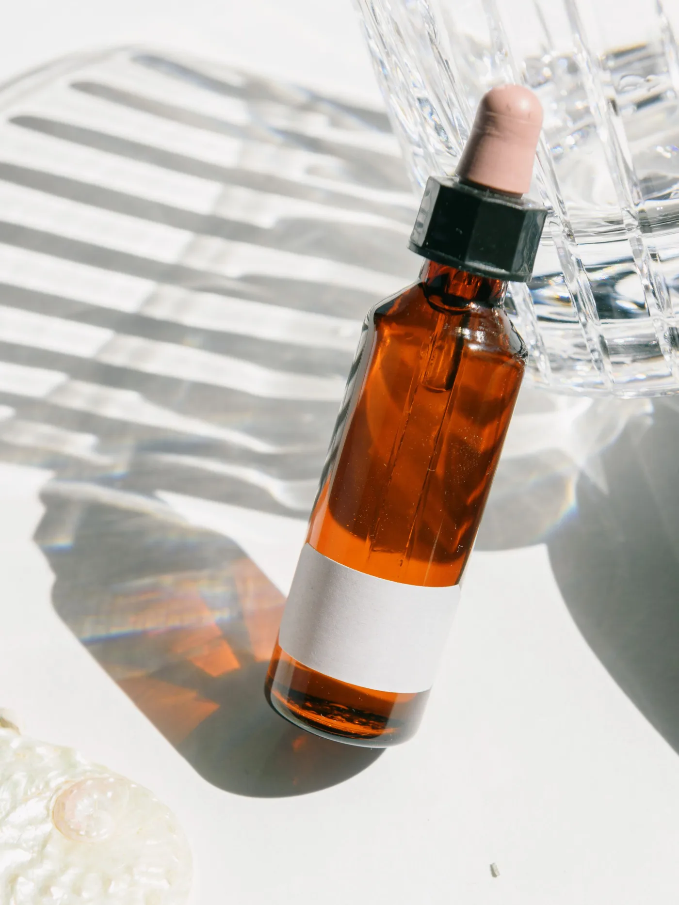 An amber dropper bottle with a blank label leaning on a glass in bright sunlight